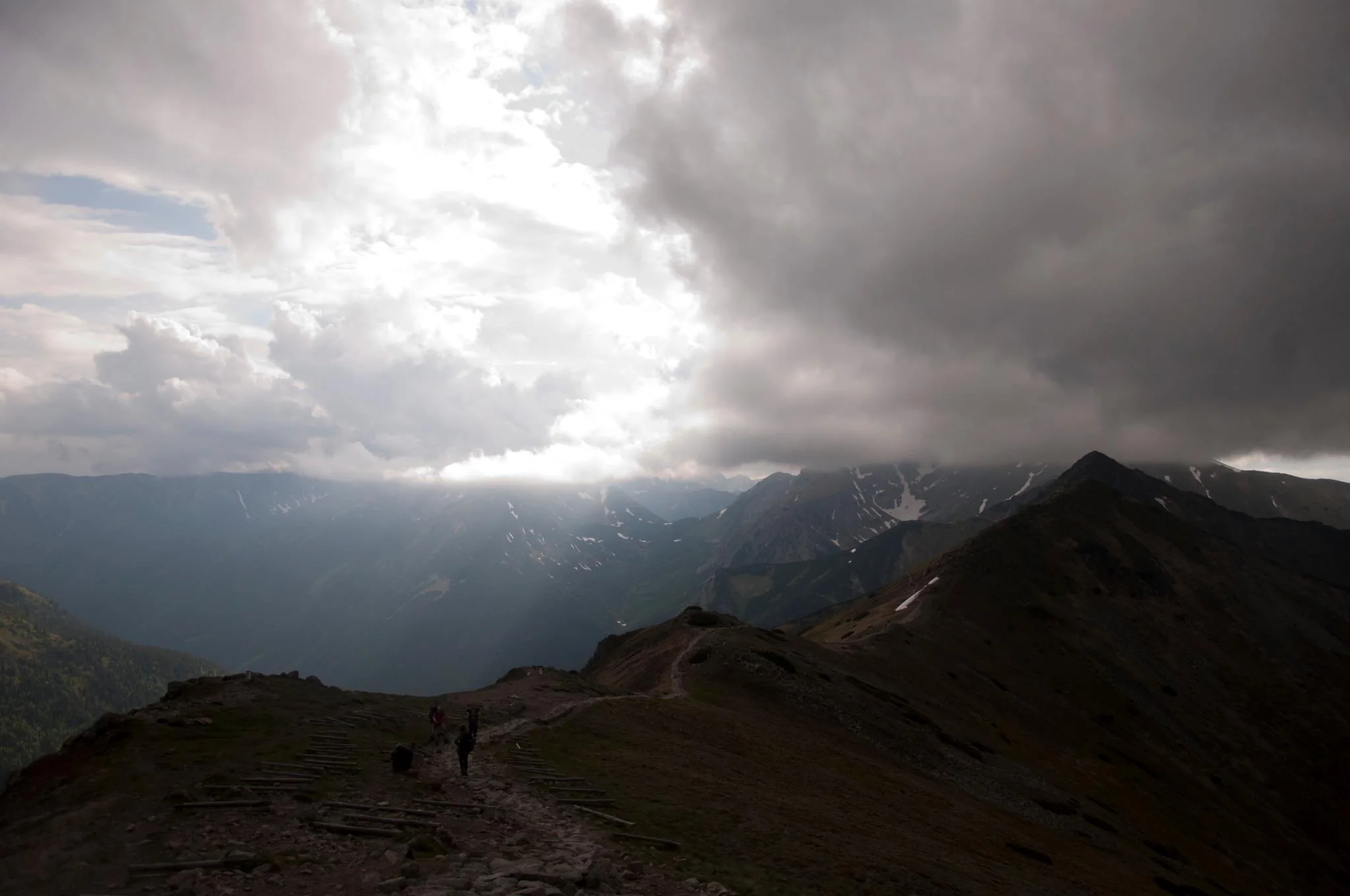 Distant hikers follow a rocky mountain ridge beneath storm clouds and a break of sunlight